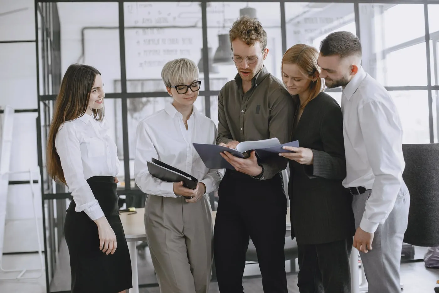 Five colleagues standing together in a bright office, reviewing documents and talking