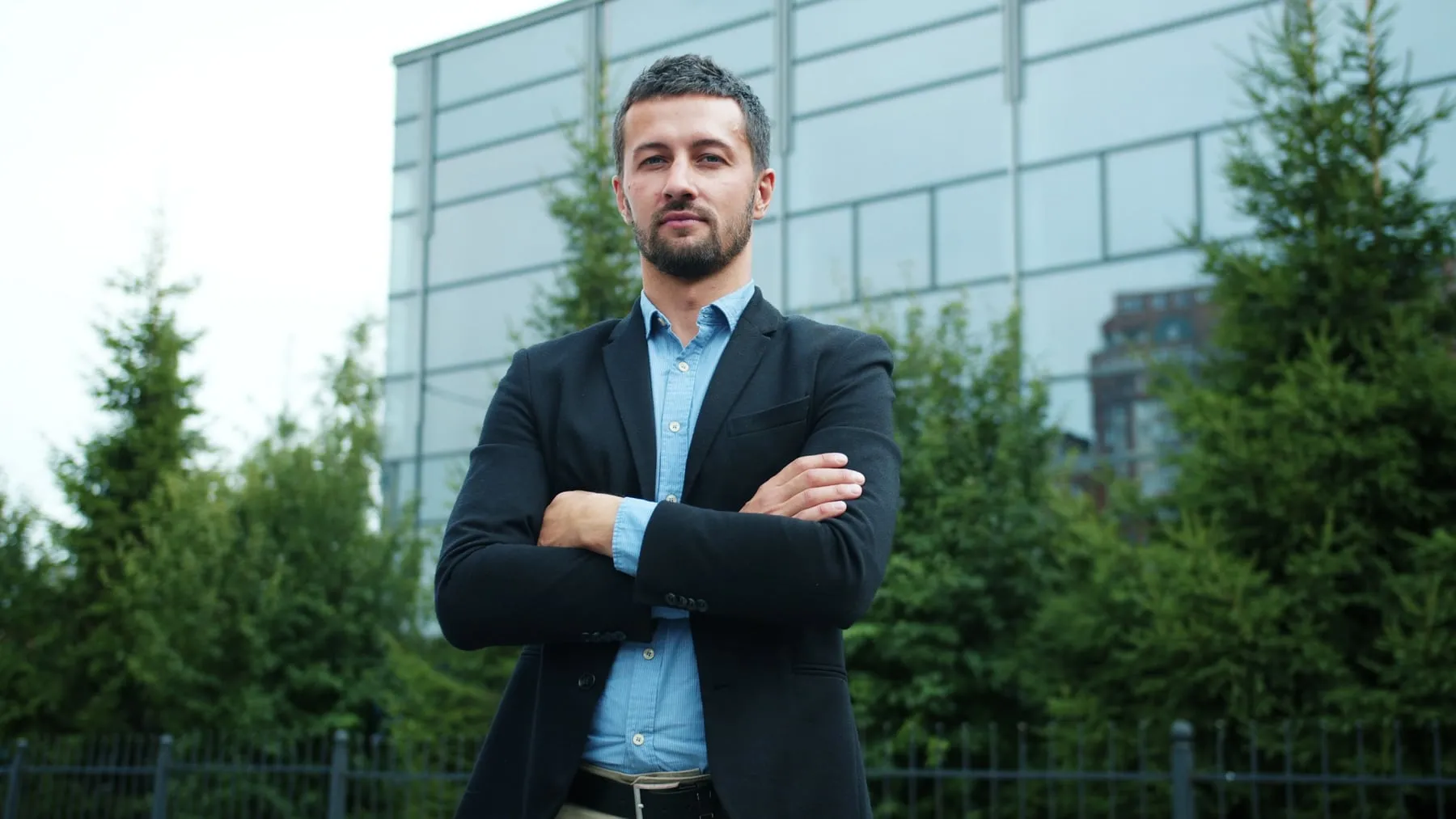 A man in a navy suit standing with his arms folded in front of an office building